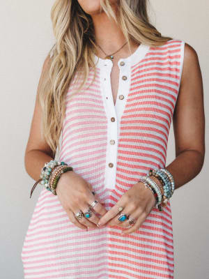 A close-up torso shot of a striped sleeveless jumpsuit in coral and white, styled with layers of beaded bracelets, stacked rings, and a delicate stone necklace — the kind of effortless summer look that feels equally at home on a boardwalk or a back p