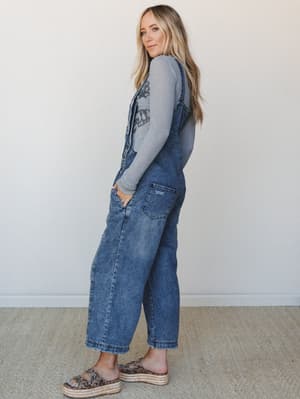 A model shot in three-quarter profile wearing slouchy wide-leg denim overalls over a long-sleeve graphic tee, styled with leopard print platform sandals — the kind of effortless, lived-in look that works just as hard for a weekend farmers market as i