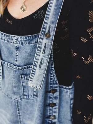 A close-up detail shot of acid-washed denim overalls layered over a black open-weave lace top, with a gold coin earring peeking into frame — the kind of effortless layering that looks intentional without trying too hard.