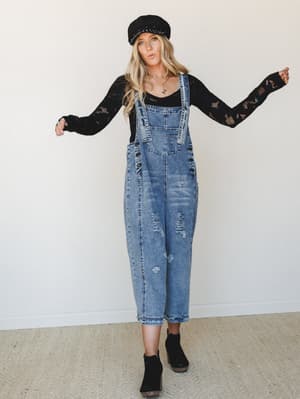 Relaxed, lived-in denim overalls styled over a black lace-knit long sleeve with ankle boots and a newsboy cap — the kind of effortless layered look that feels more 'Saturday morning with nowhere to be' than anything overthought.