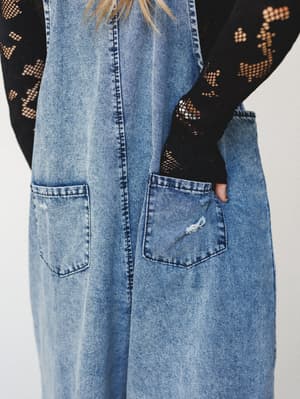 A close-up detail shot of slouchy washed denim overalls layered over a black floral crochet long-sleeve top — the kind of effortless, lived-in combo that feels like it came together without trying.