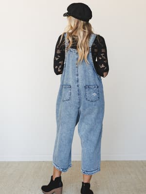 Shot from behind, a model in slouchy acid-wash denim overalls layered over a black lace-mesh top — the kind of effortless, thrown-together outfit that somehow looks completely intentional.