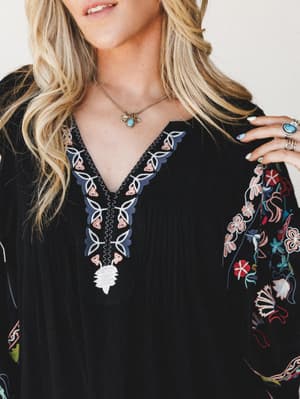 A close-up of a black dolman-sleeve top with intricate multicolor floral and botanical embroidery along the v-neckline and sleeves, styled with a turquoise stone necklace and silver rings — the kind of piece that does all the talking.