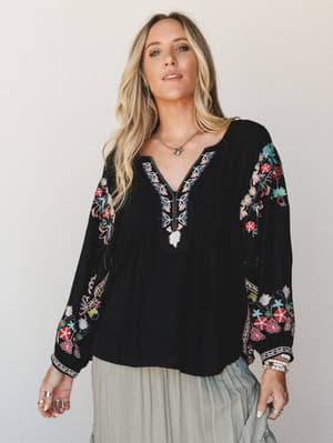 A black embroidered blouse with colorful floral sleeve detailing and a v-neckline keyhole, styled with a sage skirt and layered boho jewelry — the kind of top that does the talking for you.