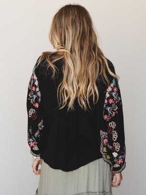 Shot from the back, this black top does all the talking — floral embroidery runs the full length of the dolman sleeves in a mix of red, blush, teal, and olive, while the solid back panel keeps it grounded and wearable.