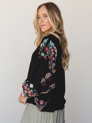 A black dolman-sleeve top covered in vibrant multicolor floral embroidery along the sleeves and hem, styled with a sage midi skirt — the kind of piece that does all the talking while you just stand there looking effortlessly put-together.