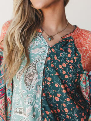 A close-up torso shot of a mixed-print top in teal, coral, and sage — three distinct floral and paisley panels pieced together with button-front closure, styled with a delicate turquoise charm necklace.