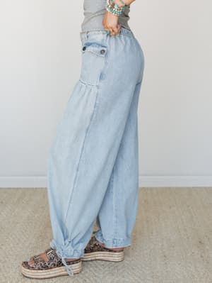 A side-profile shot of wide-leg light wash denim pants with a gathered, voluminous silhouette, styled with a gray top, leopard print platform sandals, and turquoise bead bracelets — effortless and a little unexpected.