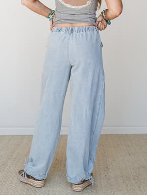 Shot from behind, these light-wash wide-leg denim pants have an easy, lived-in feel — elastic waist, ruched hem with drawstring ties, and a relaxed silhouette styled with a gray lace-trim crop top, turquoise beaded bracelets, and espadrille wedges.