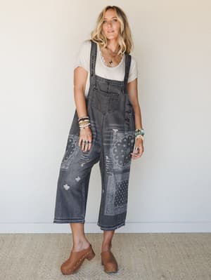 A relaxed full-body shot of a model wearing slouchy cropped denim overalls in washed black with patchwork bandana print panels and distressing, layered over a cream tee with wood-soled mules and stacked boho jewelry — effortlessly lived-in and cool.