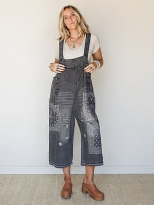 Washed black denim overalls with bandana patchwork panels and distressing, styled over a cream tee with wood clogs and layered turquoise jewelry — the kind of outfit that looks like it came together effortlessly but definitely did not.