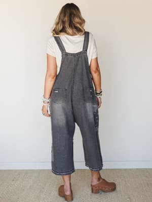 Shot from the back, a model wears slouchy acid-washed dark grey denim overalls with bandana print patchwork at the legs, layered over a cream tee and paired with brown platform clogs and stacked bracelets — effortlessly lived-in and personal.