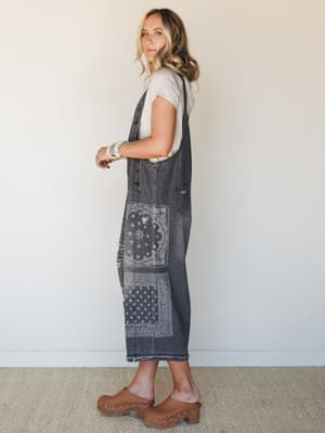 A model shot in profile wearing slouchy midi-length denim overalls in washed black with bandana patchwork panels, layered over a simple cream tee and styled with wood-studded clogs — the kind of outfit that looks effortless but has clearly been thoug