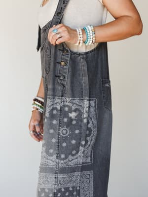 A close-up torso shot of a denim overall dress in washed black with an all-over bandana paisley print, layered over a cream tank and styled with stacks of beaded bracelets and statement rings — the kind of outfit that looks effortless but was absolut