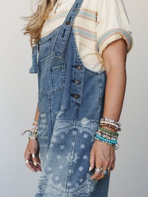 A close-up torso shot of a model wearing bandana-print denim overalls over a striped cream tee, styled with an arm stacked full of beaded bracelets and silver rings — the kind of outfit that looks like it came together effortlessly but tells you ever