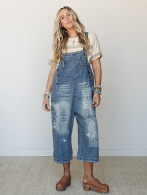 A model in slouchy cropped denim overalls with bandana-print patchwork and distressing, styled over a vintage-striped tee with brown platform clogs and stacked beaded bracelets — effortless and a little worn-in, like your coolest thrifted find.