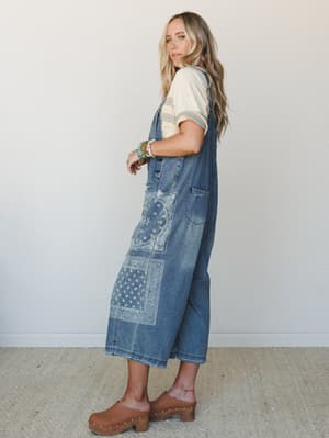 A wide-leg denim overall with bleached bandana patchwork panels is styled over a striped cream tee, finished with wood-studded clogs and a stack of colorful beaded bracelets — the kind of outfit that feels like it was thrown together perfectly.