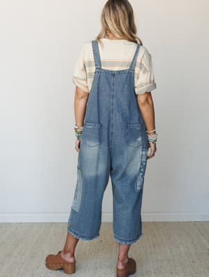 Shot from behind, a model wears wide-leg cropped denim overalls with bandana-print patchwork detailing, styled over a striped cream tee with clogs and stacked beaded bracelets — the kind of outfit that looks like it happened effortlessly but absolute