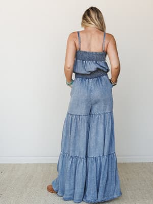Shot from behind, a model wears a flowy chambray wide-leg jumpsuit with smocked bodice and tiered leg panels — the kind of piece that looks just as good at a summer market as it does on a rooftop.