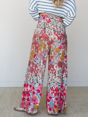 A back-view shot of wide-leg floral pants in a bold retro garden print — hot pink, marigold, and dusty blue blooms on cream — paired with a blue striped sweater for an effortlessly mixed-print moment.