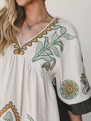 A close-up detail shot of a cream babydoll tunic with rich botanical embroidery in sage green and golden ochre — the kind of top that looks like it has a story before you even put it on.