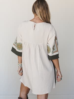 Shot from the back, this cream babydoll tunic dress does all the quiet talking — floral embroidery on the sleeves, dark contrast cuffs, and a gathered seam that says 'effortless' without trying.