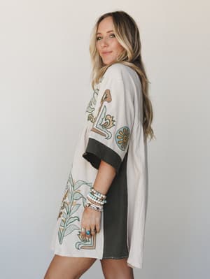 A blonde model shot in three-quarter profile wearing a cream and olive babydoll tunic dress with bold botanical embroidery, styled with a stack of beaded bracelets and turquoise rings — easy, expressive, and totally her own.