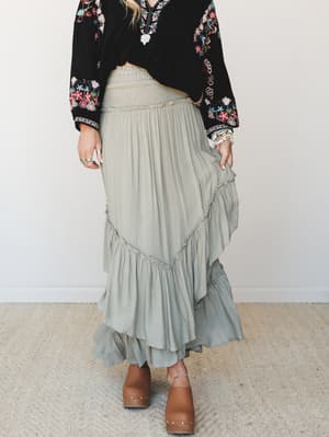 A flowy sage maxi skirt with smocked waist and layered ruffle tiers is styled with a black floral embroidered blouse and wood-heel clogs — the kind of outfit that looks like it got dressed by instinct and landed perfectly.