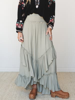 A flowy sage ruffled maxi skirt with a smocked waistband paired with a black floral embroidered top and brown clogs — earthy, expressive, and clearly at home in a weekend wandering kind of life.