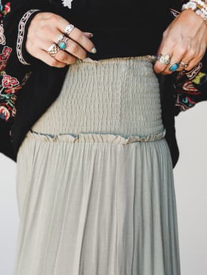 A close-up detail shot highlighting the smocked waistband and pleated skirt in a soft sage-olive, styled with a black floral embroidered top and stacked silver and turquoise rings — the kind of outfit that looks like it was put together effortlessly