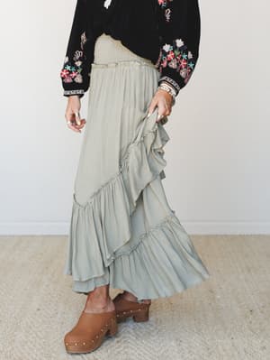 A flowy sage ruffle maxi skirt paired with a black floral embroidered top and tan studded clogs — the kind of outfit that looks effortless but was absolutely thought about.