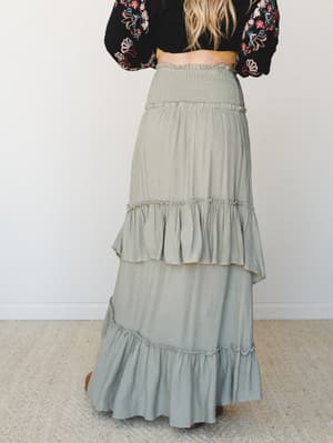 Shot from behind, a model pairs a tiered sage maxi skirt — with a smocked waist and cascading ruffle layers — against a black floral embroidered top and a mustard belt, giving off that effortlessly collected boho energy.