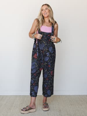 A floral black denim overall styled over a pink square-neck top with platform espadrille sandals and layered turquoise jewelry — the kind of outfit that looks effortless but was absolutely thought about.