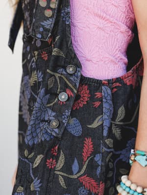 A close-up torso shot of floral black denim overalls layered over a pink textured top, showing off the botanical print, snap button closure, and a wrist stacked with turquoise and beaded bracelets — the kind of outfit that looks effortless but was de