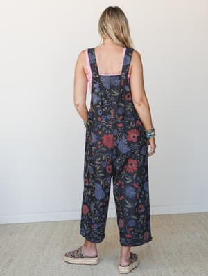 Shot from behind, a model wears slouchy wide-leg floral denim overalls in a deep black base with bold botanical print, layered over a pink tank and finished with platform espadrille sandals — the kind of outfit that looks effortless but clearly wasn'