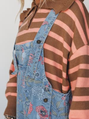A close-up detail shot layering floral-printed blue denim overalls over a brown and coral stripe knit sweater — the kind of pattern-mixing that feels effortless rather than accidental.