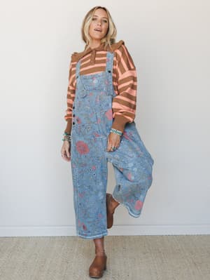 A floral-printed blue denim overall layered over a striped brown and coral sweater — the kind of outfit that looks like it came together effortlessly but someone definitely thought about it.
