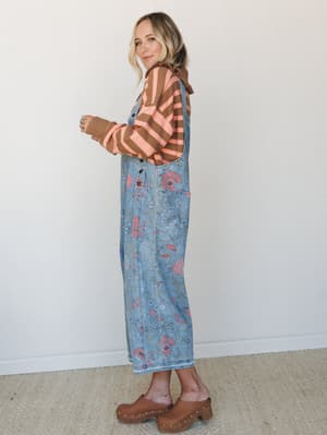 A floral-printed denim overall dress styled over a brown and coral striped long-sleeve sweater, worn with studded wood clogs — the kind of outfit that looks effortless but was absolutely thought about.