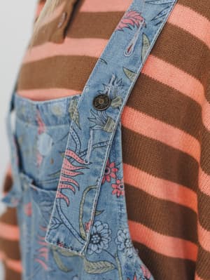 A close-up detail shot layering a floral-printed blue denim overall strap over a chunky striped knit sweater in coral and brown — the mix-and-match prints feel intentional and a little nostalgic, like something you'd put together on a good morning.