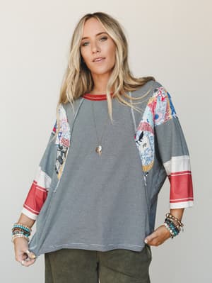 An oversized mixed-print top with fine black and white stripes, colorful patchwork panels on the sleeves and sides, and a red-trimmed neckline and cuffs — styled with olive wide-leg pants and layered boho jewelry for an effortlessly collected, lived-