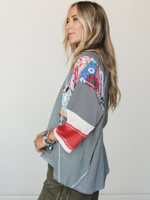 A side-profile shot of a model in a relaxed patchwork top — fine black and white stripes anchoring a sleeve full of mixed floral and bandana-print patches in red, blue, and cream — styled with olive pants and stacked boho bracelets for that perfectly