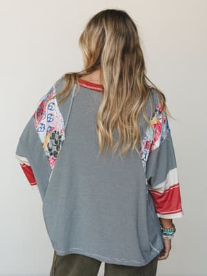 Shot from behind, a relaxed oversized top mixes fine black-and-white stripe with colorful patchwork panels at the shoulders and sleeves — the kind of top that looks like it has a past, in the best way.