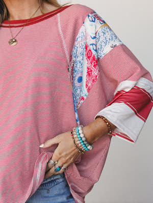 A close-up lifestyle shot of a relaxed red-and-white striped top with mixed patchwork print sleeves, styled with stacked beaded bracelets, silver rings, and a delicate pendant necklace — the kind of top that looks like it has a whole personality.