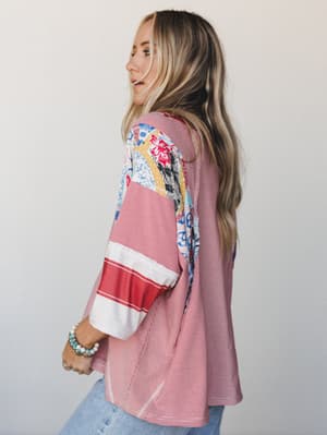 A side-profile shot of a model in a relaxed, mixed-print top — red pinstripe body meets a patchwork floral yoke and sleeve, giving it that collected, 'found-in-a-vintage-shop' energy paired with light wash jeans and stacked bracelets.