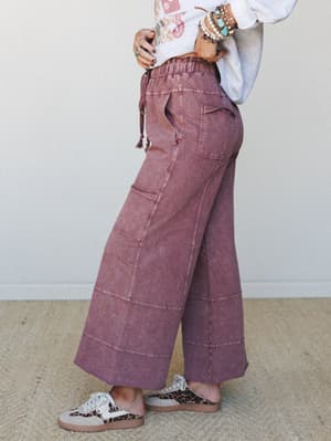 A side-angle shot of wide-leg cargo-style pants in acid-washed plum, styled with a graphic sweatshirt, stacked turquoise bracelets, and leopard sneakers — the kind of outfit that looks effortless but was definitely thought about.