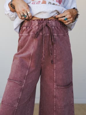 A close-up mid-shot of wide-leg cargo-style pants in a washed plum tone, styled with a graphic sweatshirt and layered silver and turquoise rings — the kind of effortless pairing that looks intentional without trying.