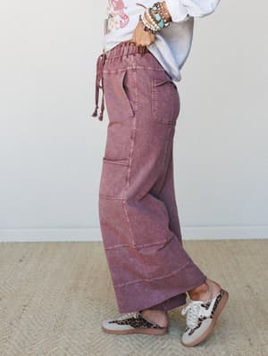 A side-profile shot of wide-leg cargo pants in a washed plum colorway, styled with a graphic sweatshirt, leopard print sneakers, and a stack of mixed bracelets — casual and collected with just enough personality.