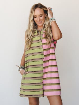 A color-blocked striped mini dress with a relaxed A-line silhouette — split down the center with lime green and pink stripe panels — styled with stacked boho bracelets and a laid-back smile that feels like the first day of a road trip.