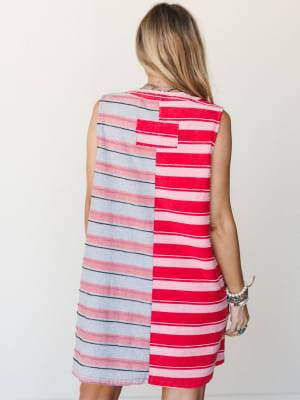 A colorblock stripe mini dress shot from the back, where two mismatched stripe panels — one red and pink, one grey and navy — come together in a playful, intentional patchwork feel, styled with layered boho jewelry.