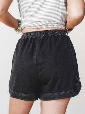 A back-view shot of faded black lounge shorts styled with a striped crop top and stacked beaded bracelets — the kind of effortless, lived-in look you actually want to wear every day.